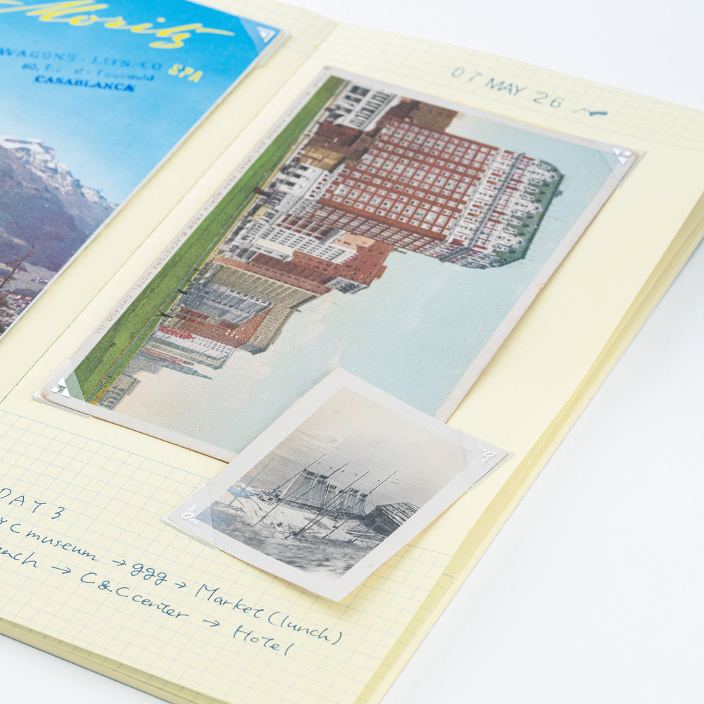 A vintage-style travel postcard album page with handwritten notes, featuring a large tilted postcard of a red-roofed building and a smaller black-and-white postcard of ships, alongside a partial blue postcard in the corner.