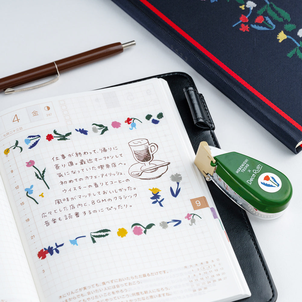 An open planner or notebook with Japanese writing and colorful floral deco rush tape, styled on a white surface with a pen, a green tape dispenser, and a dark patterned notebook.