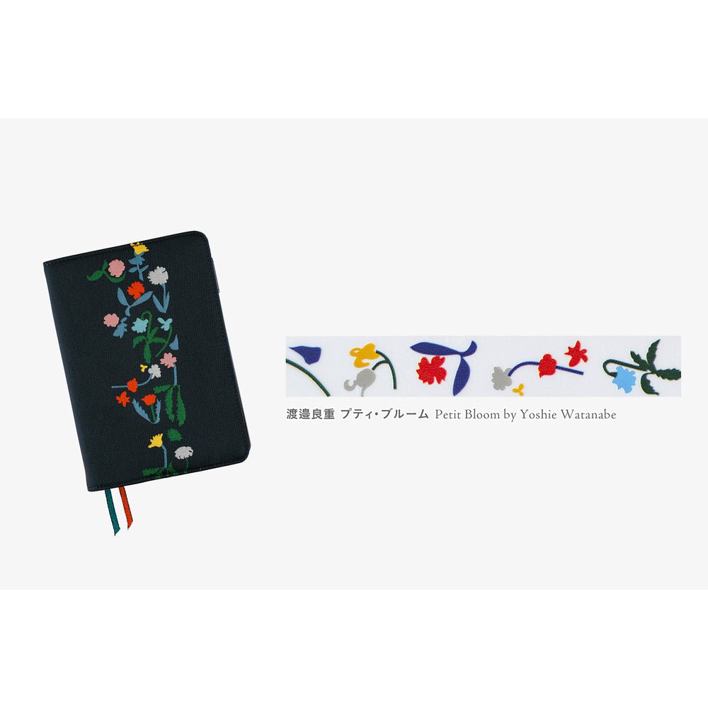 A black notebook cover with colorful floral embroidery on the front, shown on a light gray background. To the right is a close-up strip of floral deco rush tape with Japanese text and 'Petit Bloom by Yoshie Watanabe'.