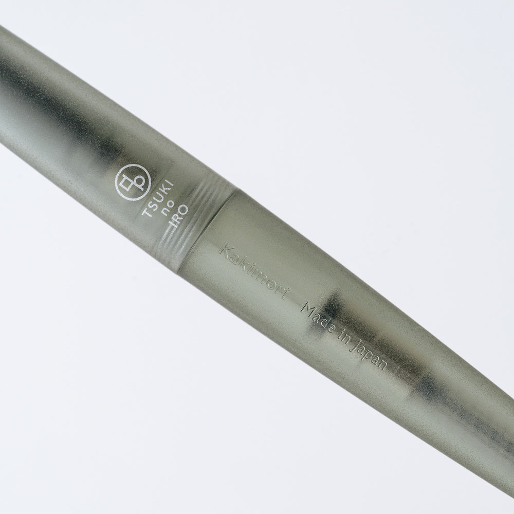 A close-up of a green-gray TSUKI no IRO rollerball pen with engraved branding and text, set against a clean white background.