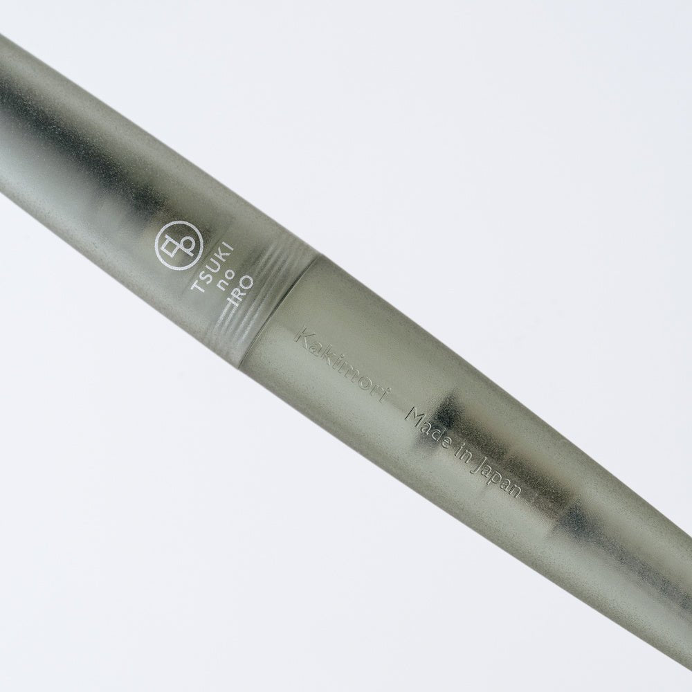 A close-up of a green-gray TSUKI no IRO rollerball pen with engraved branding and text, set against a clean white background.