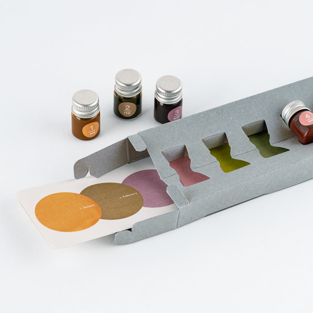 A gray cardboard tray holding four small glass ink bottles with silver caps, shown on a clean white background. A printed card with circular color swatches is partially slid out from the tray.