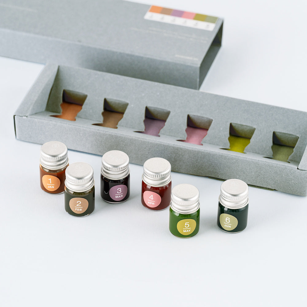 Six small glass bottles with silver caps, each containing a different colored ink, arranged in front of an open gray box with matching slots. The bottles are labeled 1 through 6 with circular stickers.