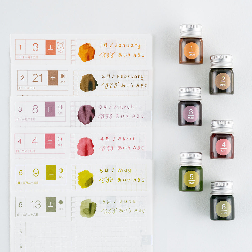 Six small glass bottles of ink with silver caps are arranged next to a white monthly planner page showing color swatches and handwritten notes in matching shades.