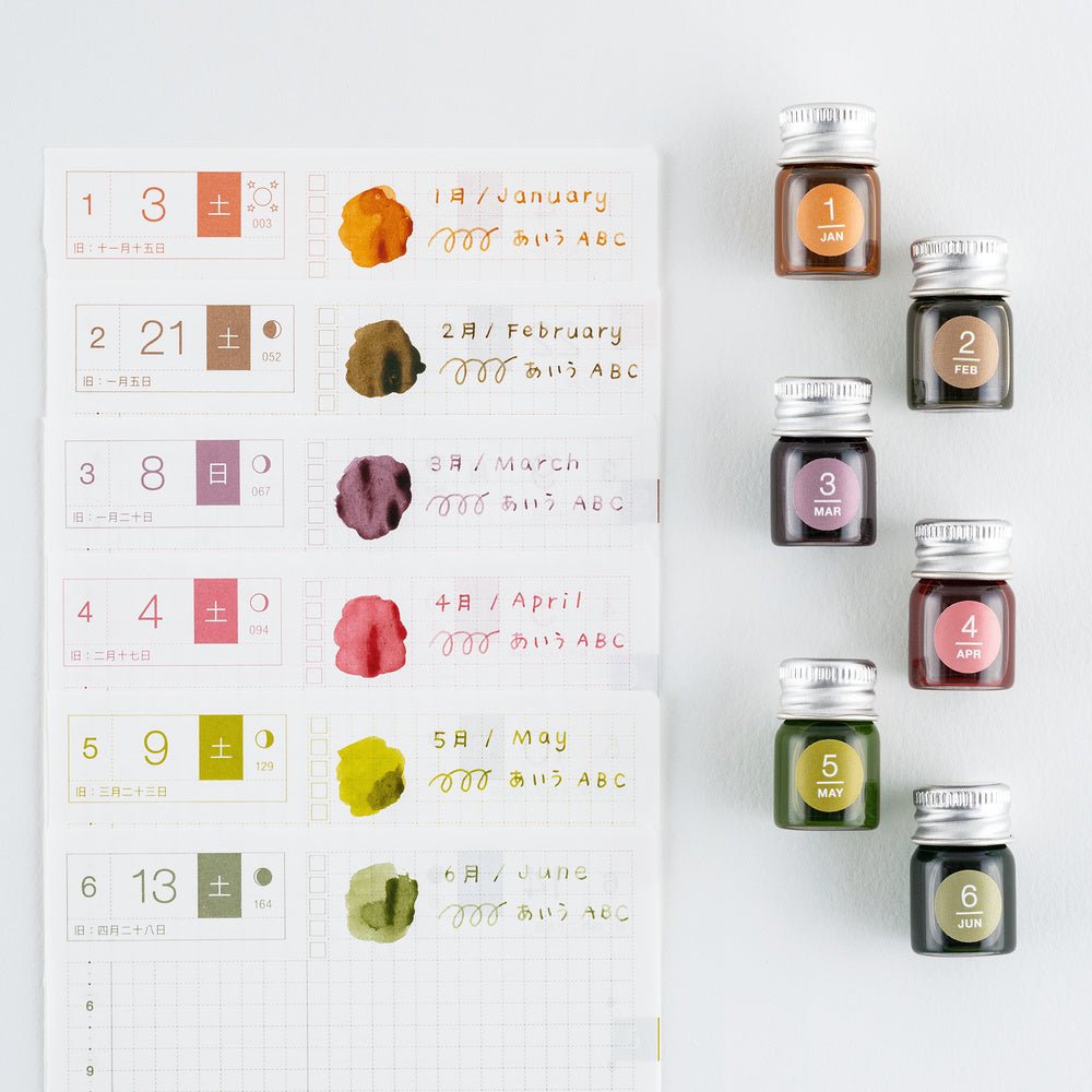 Six small glass bottles of ink with silver caps are arranged next to a white monthly planner page showing color swatches and handwritten notes in matching shades.