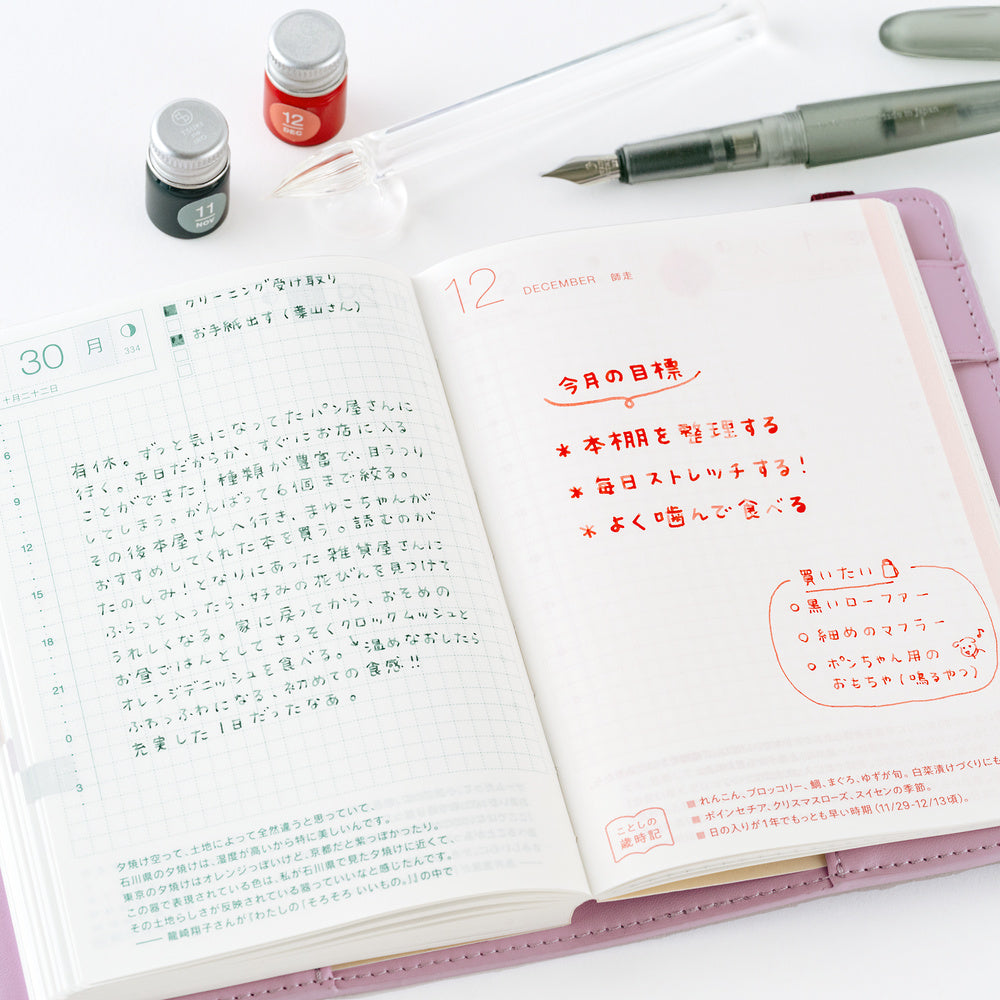 An open pink planner showing handwritten and printed Japanese notes, photographed on a white surface with a pen, two small bottles, and a glass quill nearby.