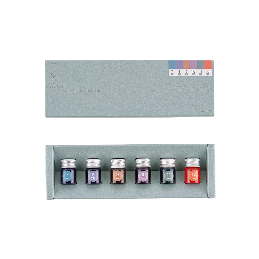 A set of six small glass ink bottles with silver caps arranged in a light gray presentation box, shown below the matching box lid. The bottles have color labels numbered 7 through 12.