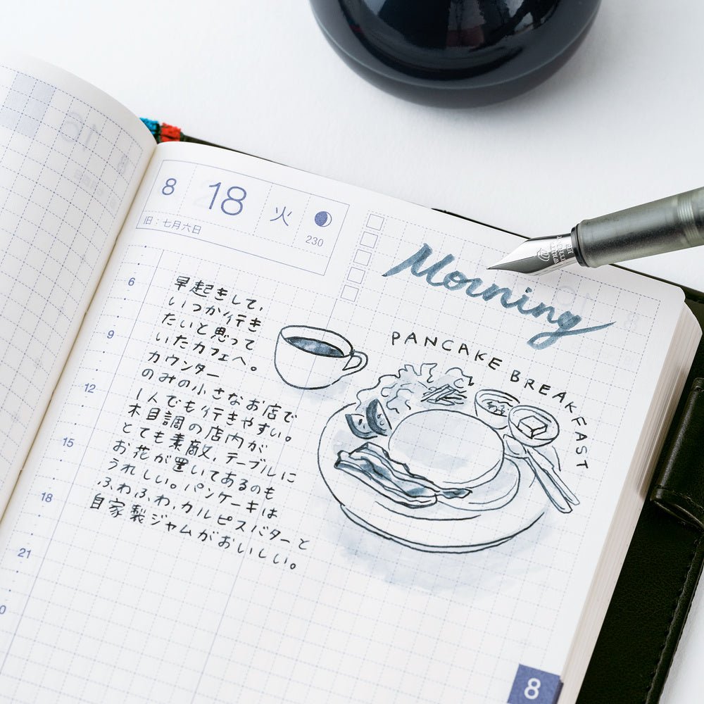 An open grid planner or journal showing a decorated morning log with handwritten text, a coffee cup sketch, and the words 'Morning Pancake Breakfast.' A silver pen rests on the page, and part of a dark round object is visible at the top against a white background.