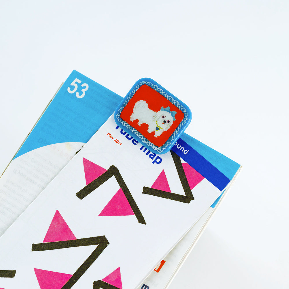A close-up of a colorful paper clip with a blue border and a red square featuring a white dog, placed on top of a printed page.