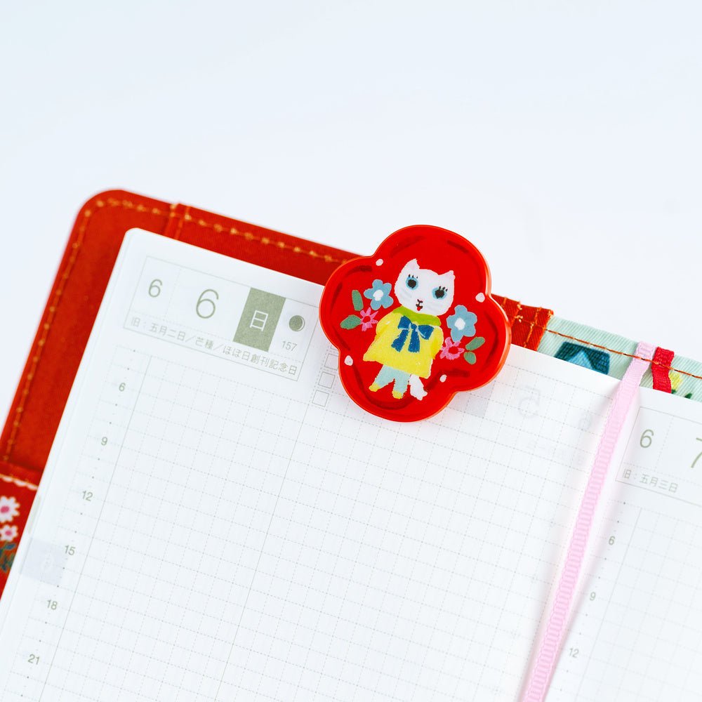 A red floral planner clip with a cartoon cat character in a yellow dress, holding an open grid planner with a pink ribbon bookmark.