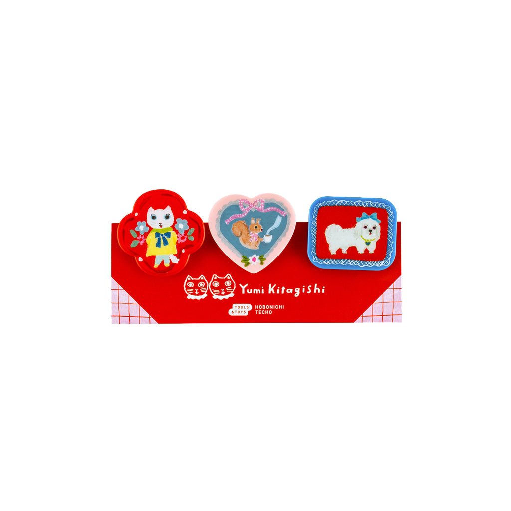 Three Yumi Kitagishi-themed clips displayed on their red branded card packaging against a solid white background. The pins include a round pin with a red cat, a heart-shaped pin with a chipmunk, and a square pin with a white dog.