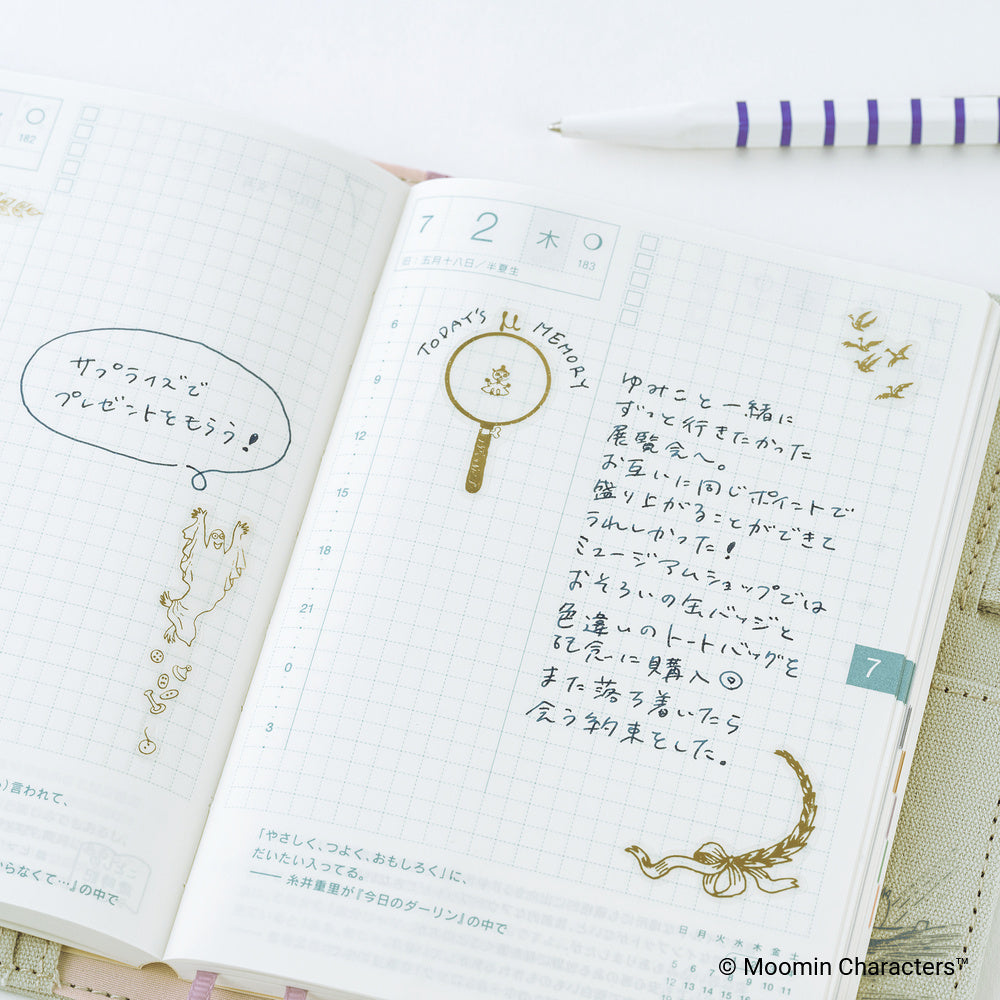 An open planner with Japanese text, grid pages, decorative doodles, gold stickers, and a purple-and-white striped pen on a light surface.
