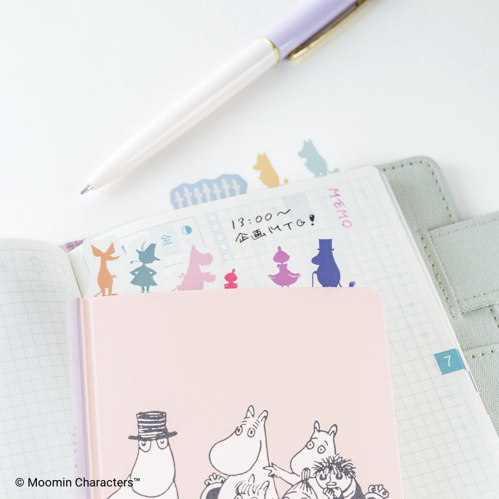 A pink Moomin Character notebook with black-and-white line art of characters and colorful silhouette index tabs, placed on top of an open grid planner. A white pen with a lavender and gold cap rests diagonally across the scene.