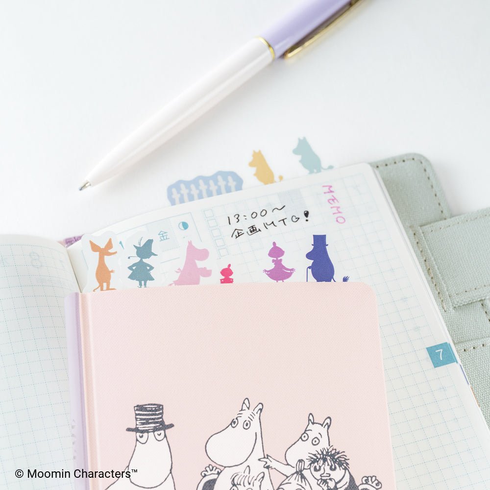 A pink Moomin Character notebook with black-and-white line art of characters and colorful silhouette index tabs, placed on top of an open grid planner. A white pen with a lavender and gold cap rests diagonally across the scene.
