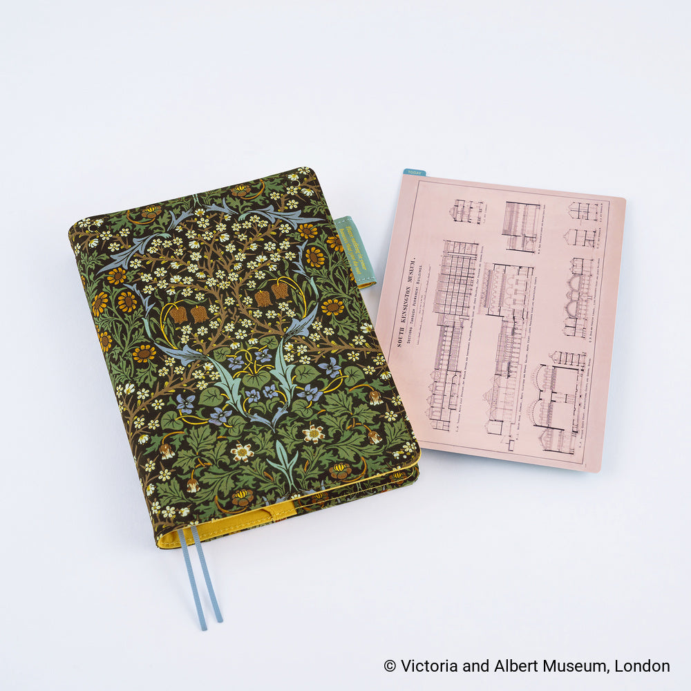 A decorative notebook with a floral and tree pattern in green, yellow, and blue, shown closed with a ribbon bookmark. Next to it is an A5 pencil board with architectural line drawings on a pale pink background.