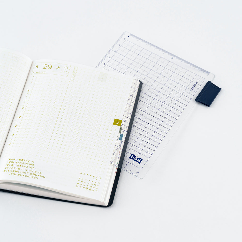 An open planner with a grid page and gold page marker, placed on top of a clear pencil board with a dark blue corner piece and gridded design, on a clean white background.