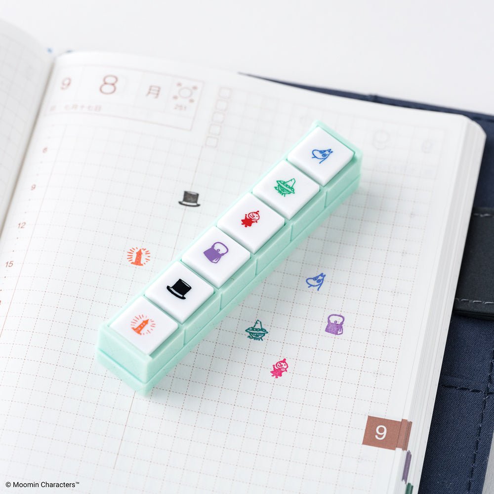 A mint green Pochitto6 push-style button stamp with six Moomin designs against a gridded notebook page