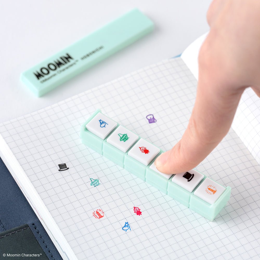 A finger presses a mint green Moomin-themed Pochitto6 stamp on a graph notebook. The pad has six white square buttons with colorful cartoon character icons, and the Moomin brand is visible on the matching mint green case in the background.