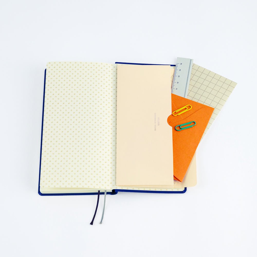 An open Hobonichi Techo Weeks planner with a cream paper pocket envelope open with stationery supplies on white background