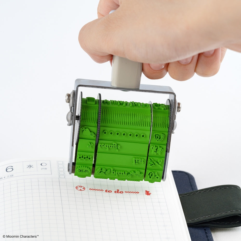 A hand holding a green rubber rotating stamp over a lined notebook, showing the stamped characters and text.