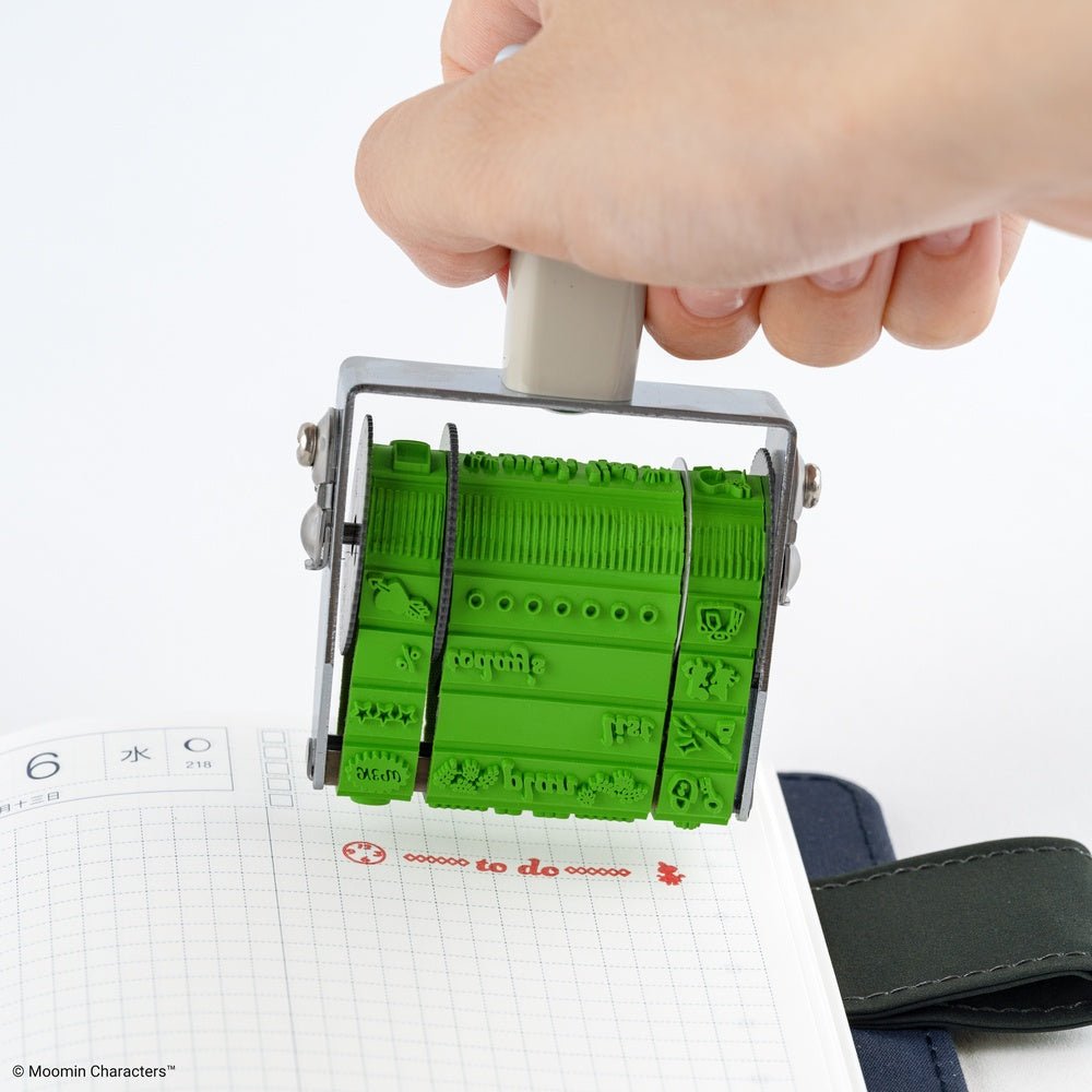 A hand holding a green rubber rotating stamp over a lined notebook, showing the stamped characters and text.