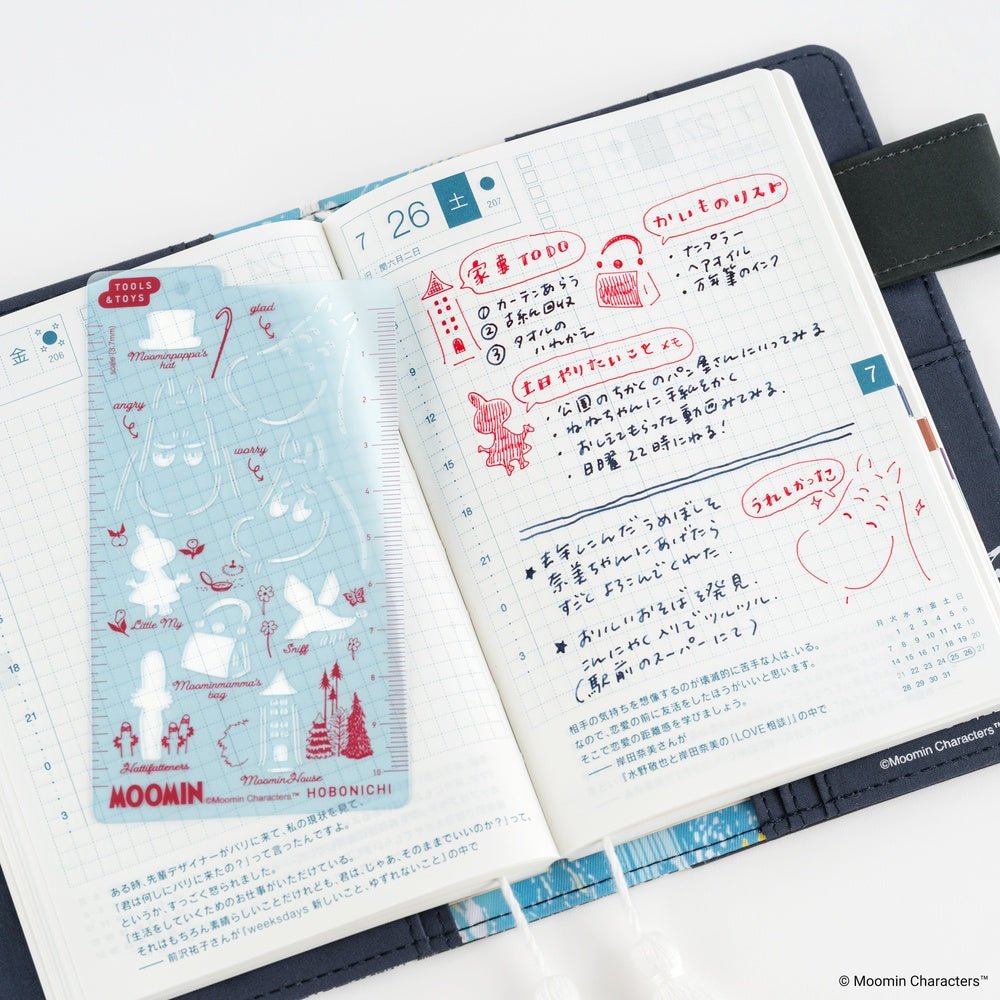 An open planner showing decorated pages with Japanese writing and illustrations inspired by Moomin, including a light blue stencil photographed on a white background.