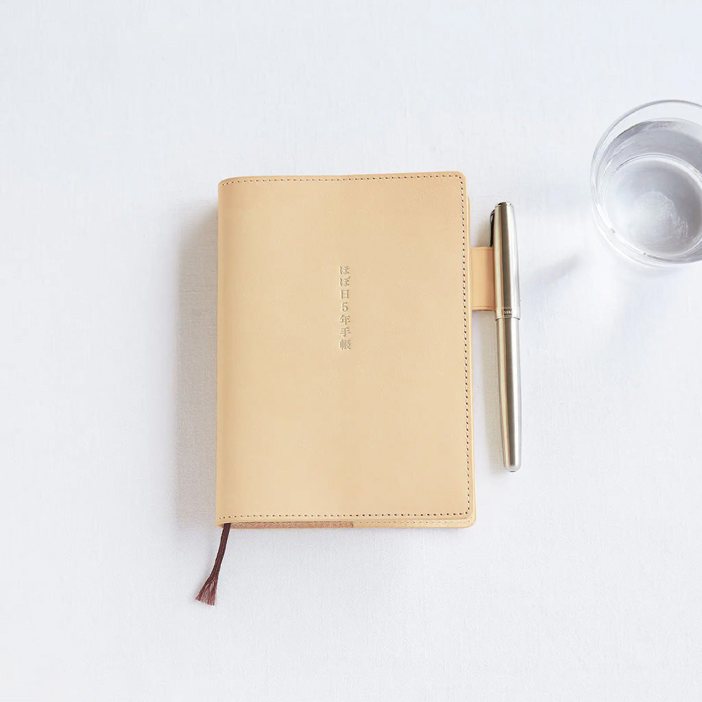 A natural beige leather notebook cover for Hobonichi 5-Year Techo next to pen on white background