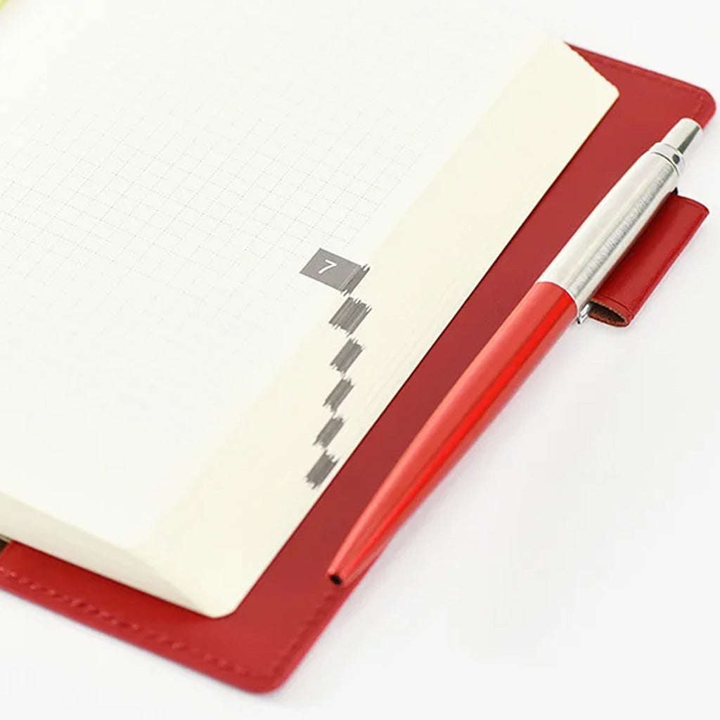 A red leather planner cover holding an open cream-colored grid notebook, with a matching red and silver pen resting on the cover, set against a clean white background.