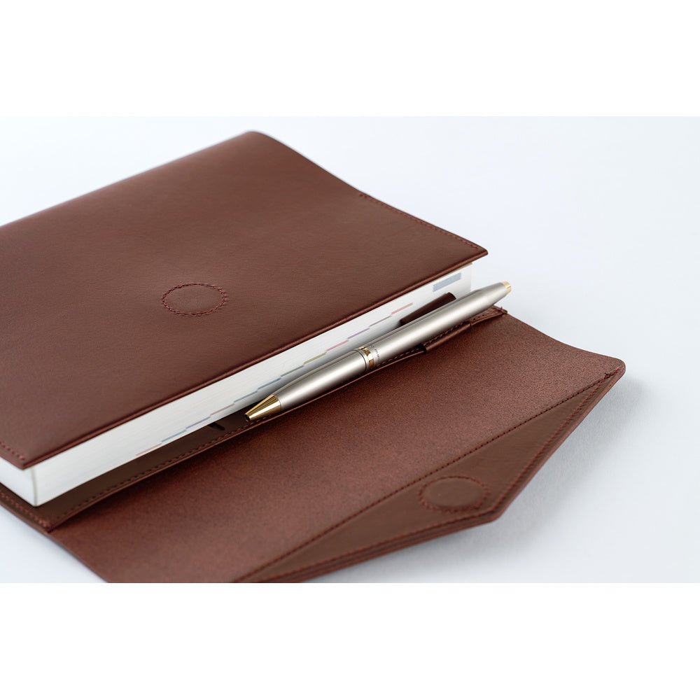 A brown leather Hobonichi Techo cover holding a white planner and a pen in a pen loop, photographed on a white background.