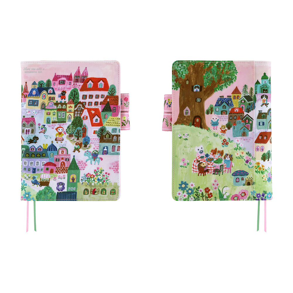 Two illustrated notebook covers shown side by side on a plain white background. Both sides are light pink and show a whimsical village scene featuring colorful houses, trees, and small animal figures.