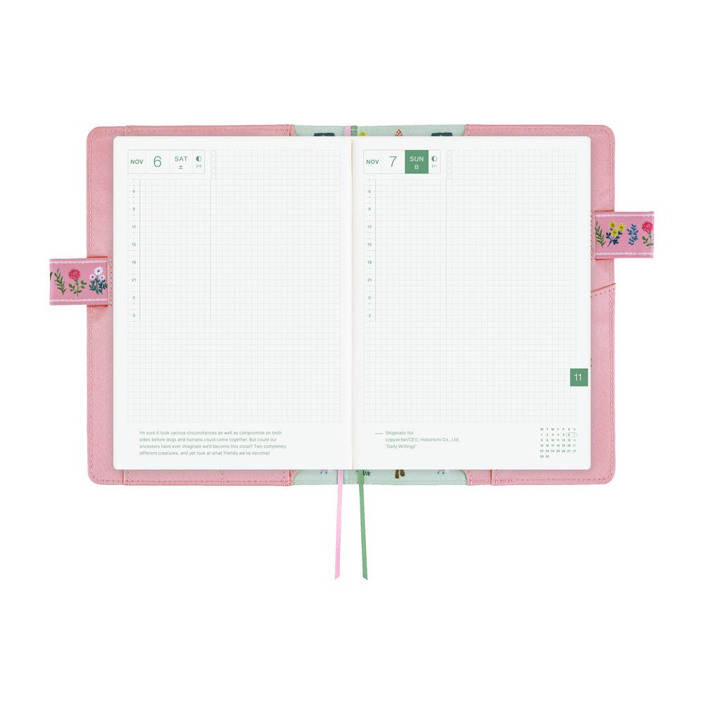 An open planner with a pink cover and white grid pages, shown on a plain white background. The cover has small floral details on the side tabs, and two ribbon bookmarks in pink and green hang from the bottom.