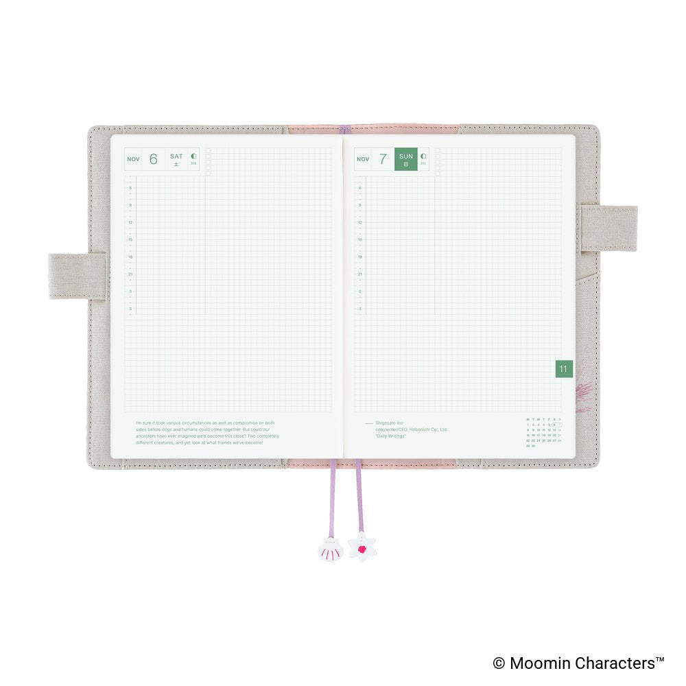 An open monthly planner with two grid pages visible, a beige cover, pink and lavender ribbon bookmarks, and decorative charms, shown on a white background.