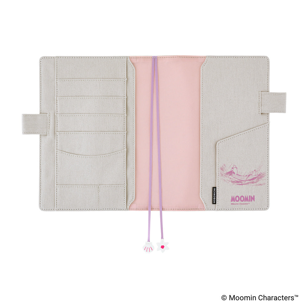 An open Moomin planner cover in a light beige cotton material with a pink fabric accent. It features multiple card slots, pen loops, and purple ribbon bookmarks with small charms at the ends.