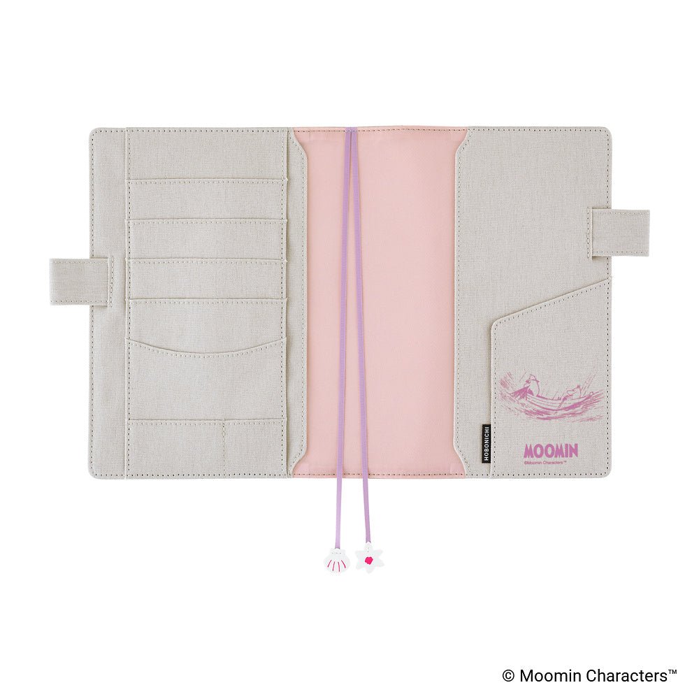 An open Moomin planner cover in a light beige cotton material with a pink fabric accent. It features multiple card slots, pen loops, and purple ribbon bookmarks with small charms at the ends.