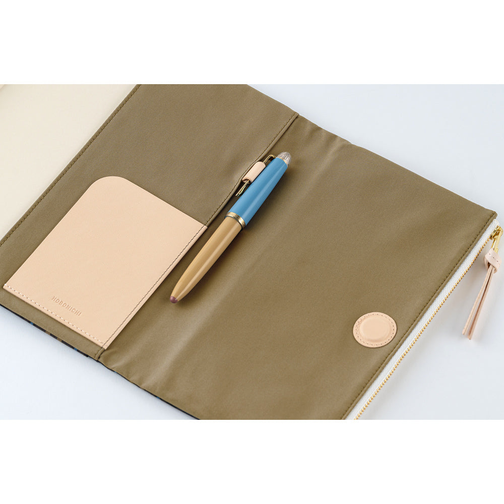 An open beige planner cover with a natural leather pocket, a blue and gold pen, and a gold zipper pull, photographed on a white background.