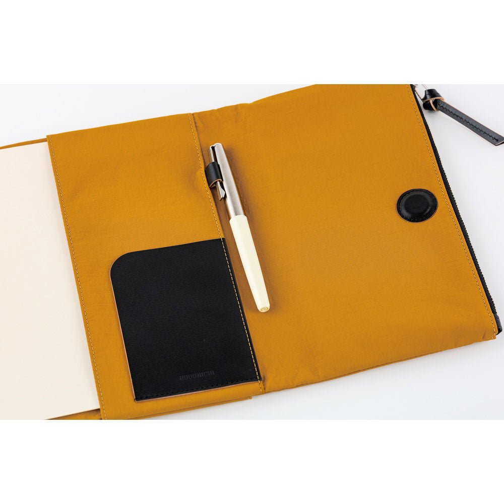 Close-up of the interior of a mustard yellow Hobonichi Tragen cover, showing a black pocket, pen holder with pen, and magnetic closure on a white background.