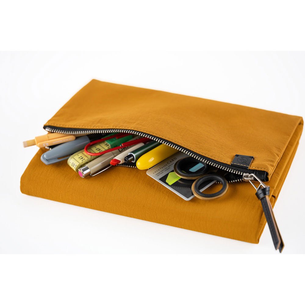 An open zippered pouch on a mustard yellow Hobonichi Tragen cover filled with pens, scissors, tape, and other stationery accessories on a white background.