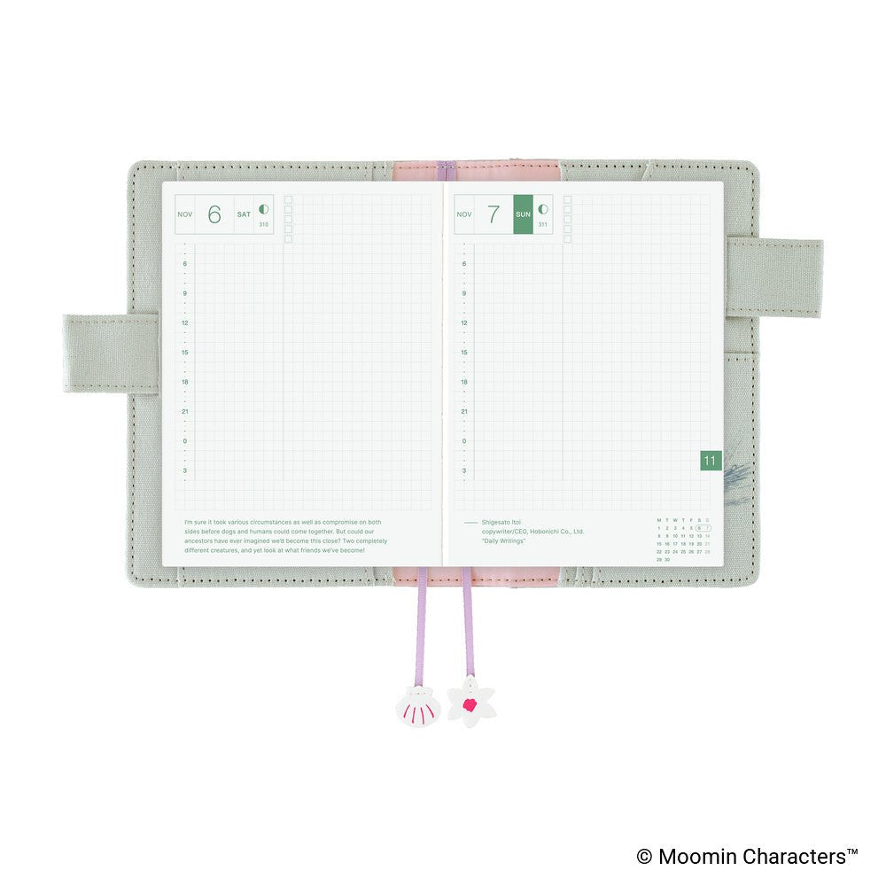 An open planner with two grid pages visible, a pale sage green cover, lavender ribbon bookmarks, and decorative charms, shown on a white background.