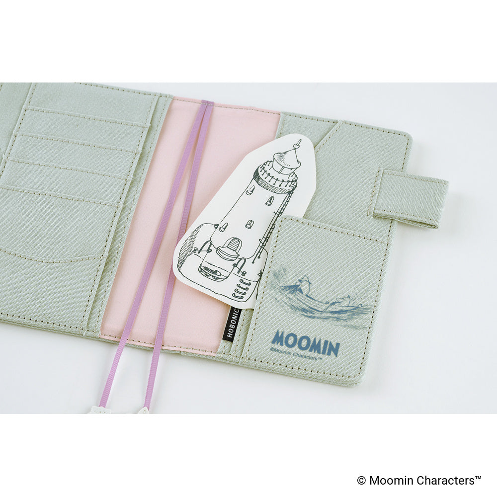 A pale sage green faux-leather card holder wallet with a pink elastic band, featuring a white lighthouse-shaped card with a black line drawing and the blue 'MOOMIN' logo.