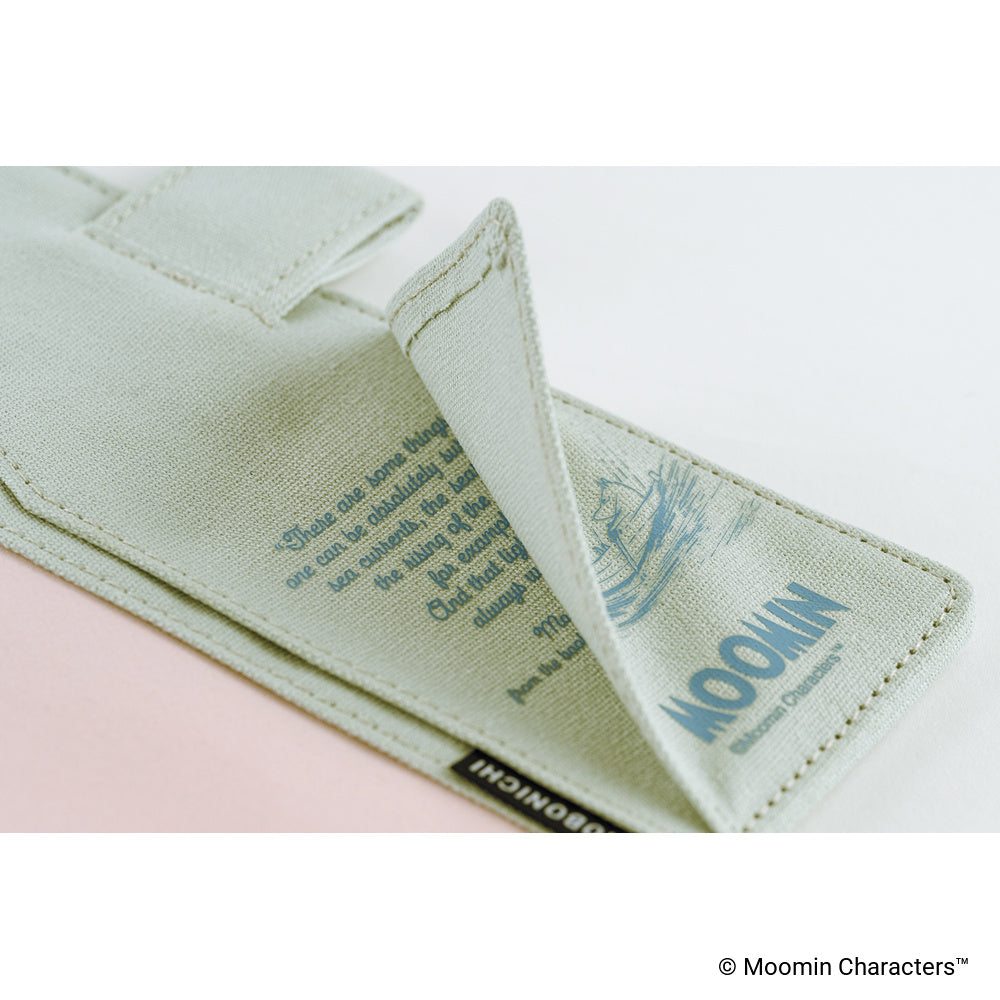 A close-up of a pale sage green fabric accessory, likely a pouch or strap, with stitched edges, printed blue text and illustration, and a small black brand tab reading Moomin. The fabric is photographed on a clean white background with a soft pink area visible underneath.