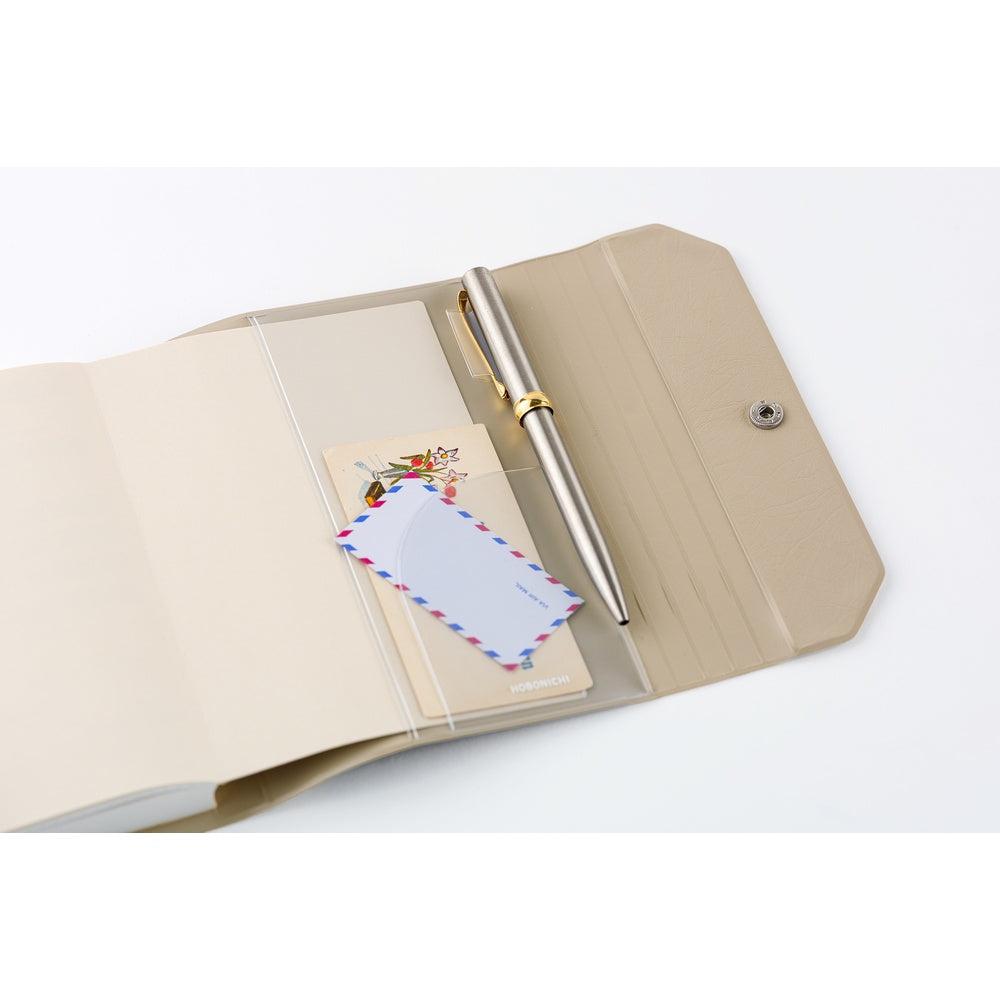 A beige notebook holder shown open on a white background, containing a clear plastic pocket with a silver pen and a postcard.