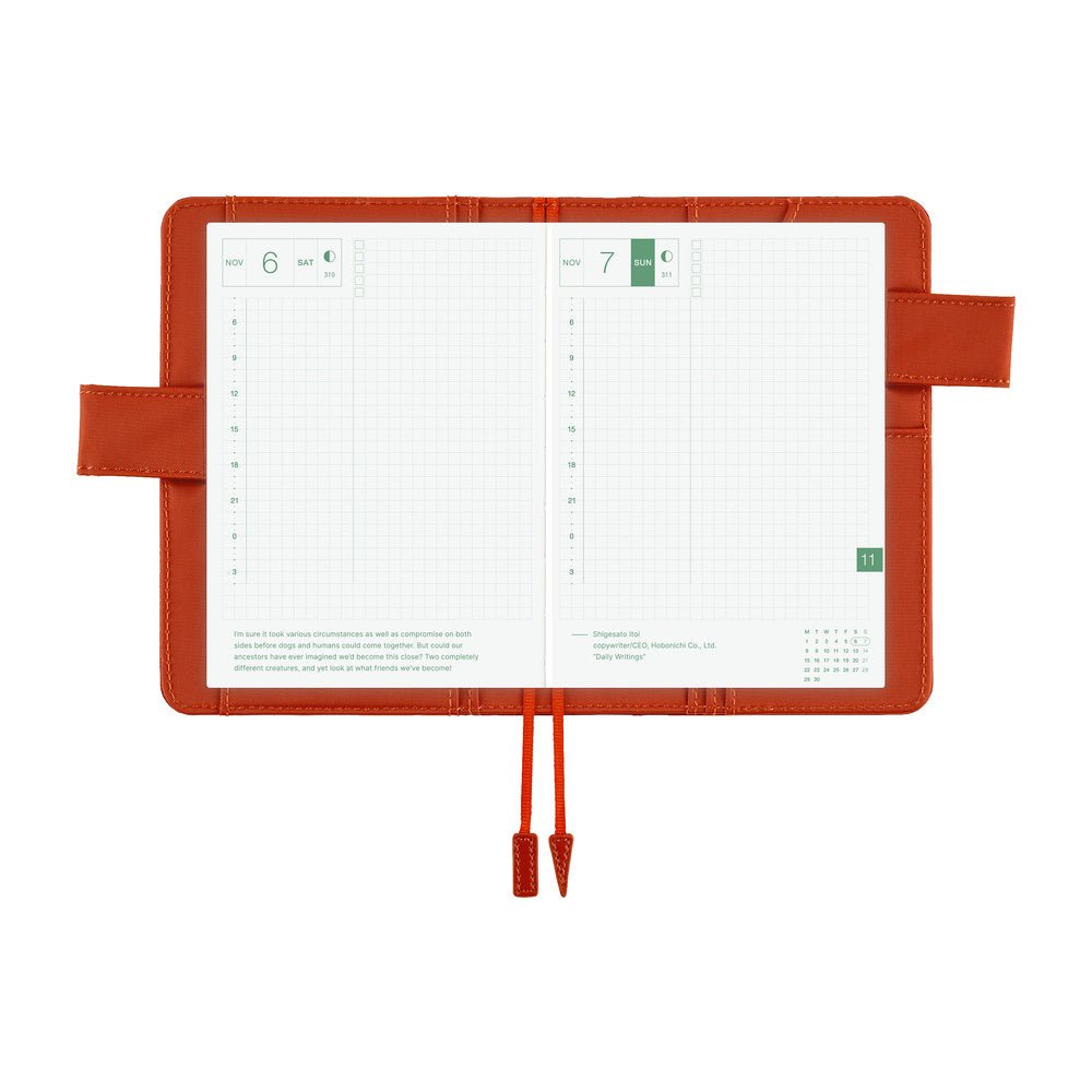 An open grid planner with a reddish-orange cover, showing two white daily pages with green date markers and printed text, photographed on a plain white background.