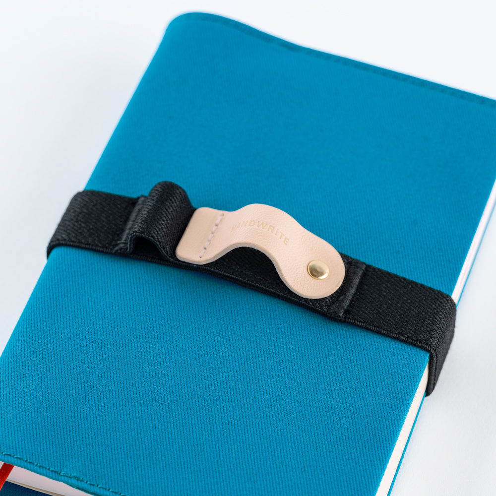 A close-up of a blue notebook or journal secured with a black elastic strap and a beige leather buckle. The buckle has the brand name 'Handwrite' embossed on it.