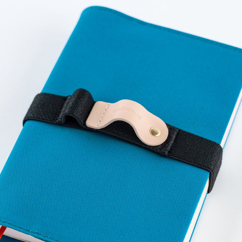 A close-up of a blue notebook secured with a black elastic strap and a beige leather pen holder. The leather has the brand name 'Handwrite' embossed on it.