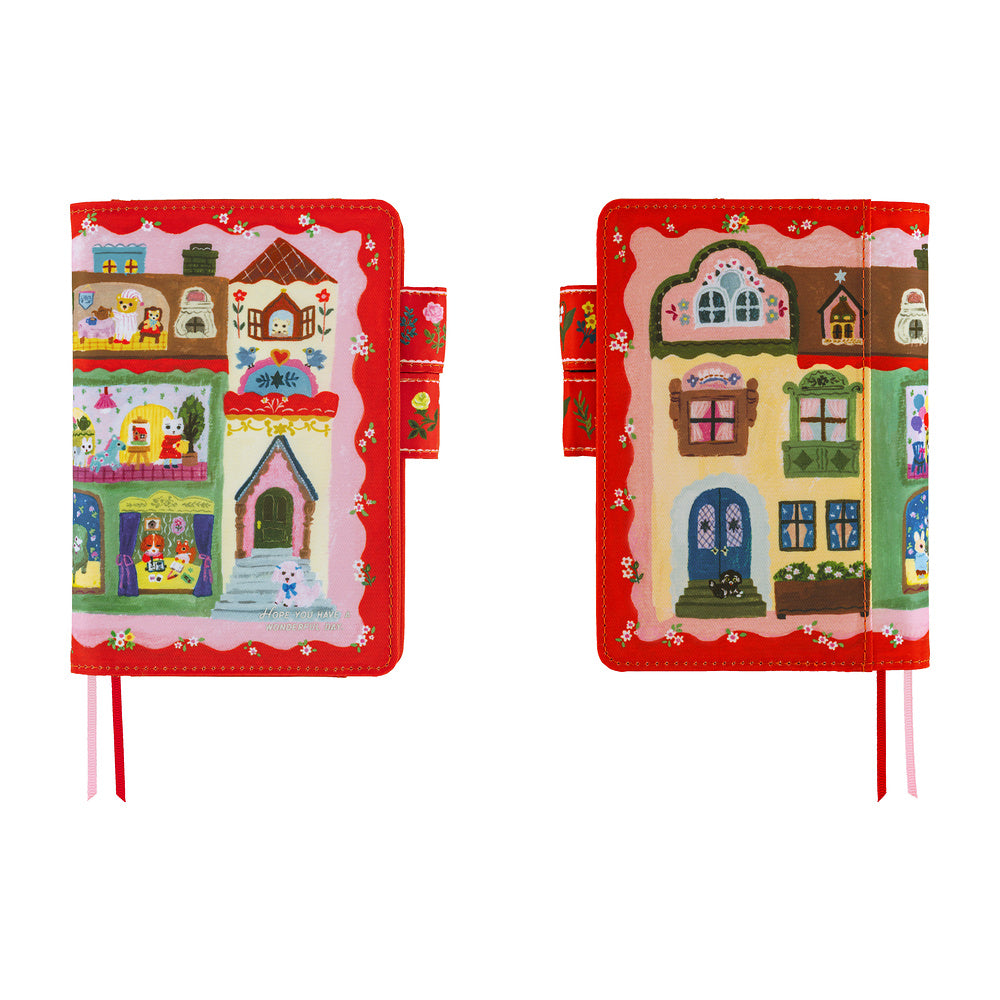 Two views of a small fabric journal or notebook cover with a colorful whimsical house illustration on a red border, shown on a plain white background.