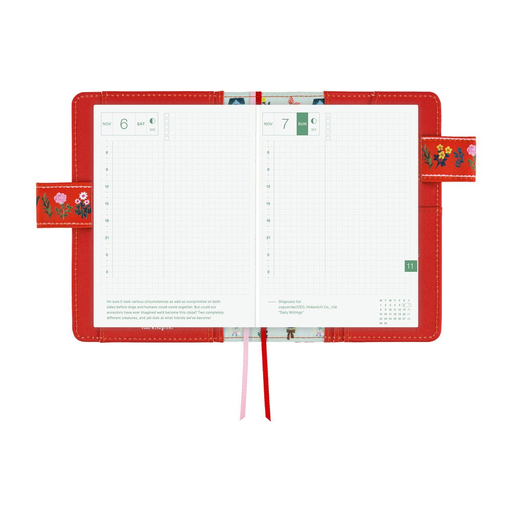 An open red notebook cover with planner grid pages, floral details, and two ribbon bookmarks, photographed on a clean white background.