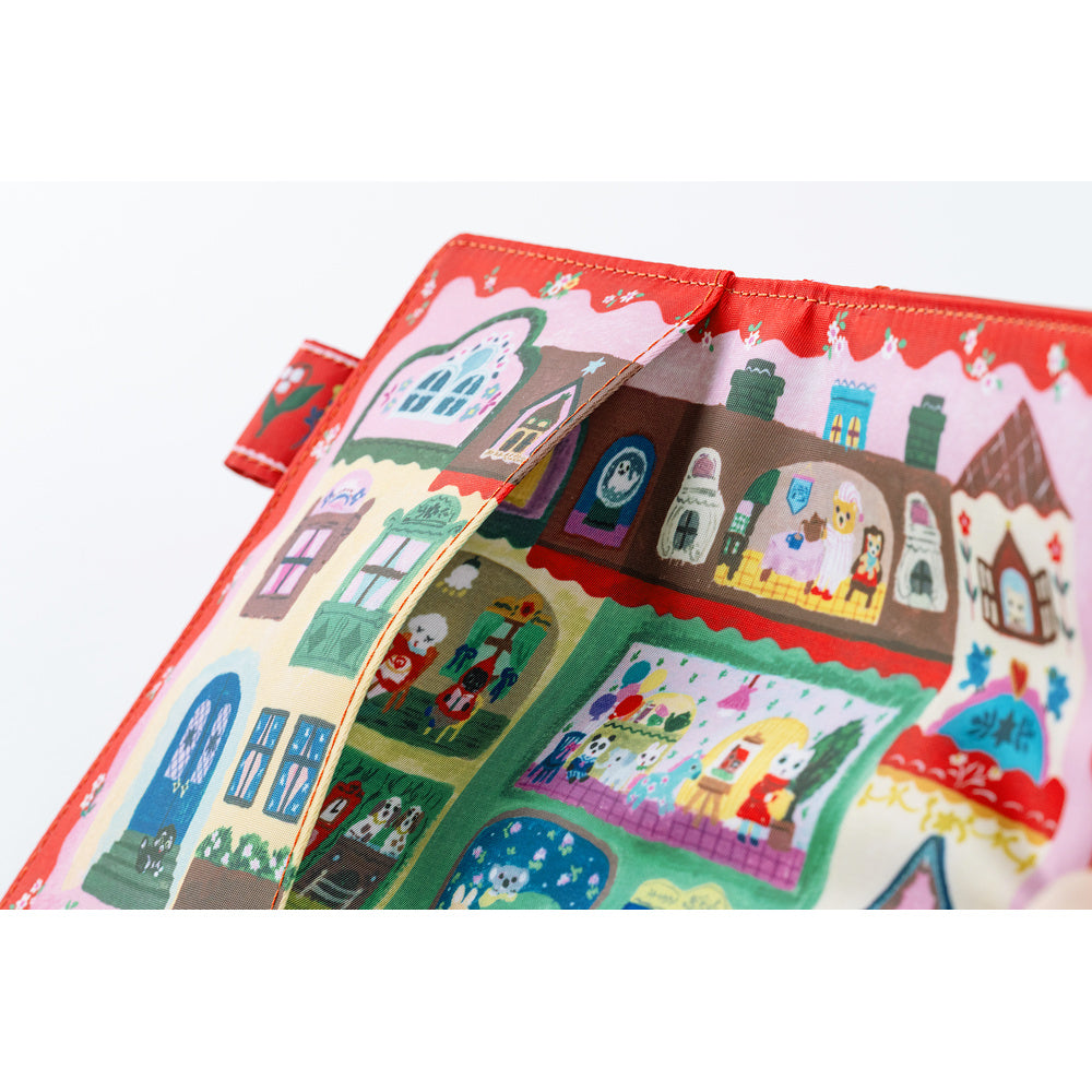 A close-up of a fabric accessory, likely a pouch or bag, featuring a colorful winter village illustration with houses, characters, and snowy scenes. The fabric has a red stitched border and a small matching tab on the left side, photographed against a plain white background.