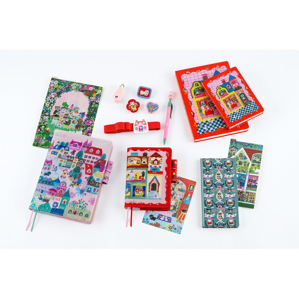 A flat lay of colorful stationery and accessories on a white background, including illustrated notebooks, sticker sheets, a pink pen, small charms, and a red hair tie.