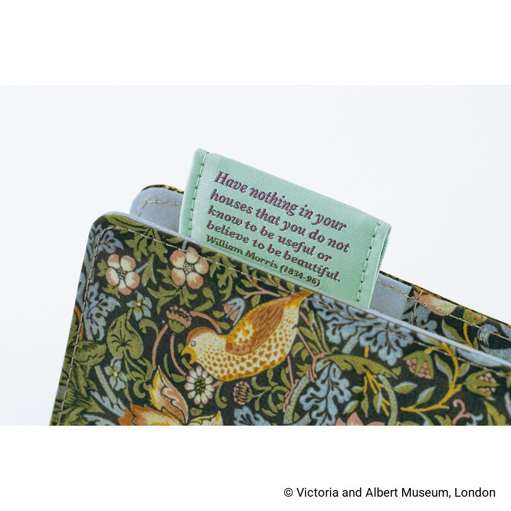 Close-up of a book with a decorative cover featuring a William Morris-style bird and floral pattern, with a pale green fabric bookmark or label partially pulled out. The label contains a quote by William Morris.
