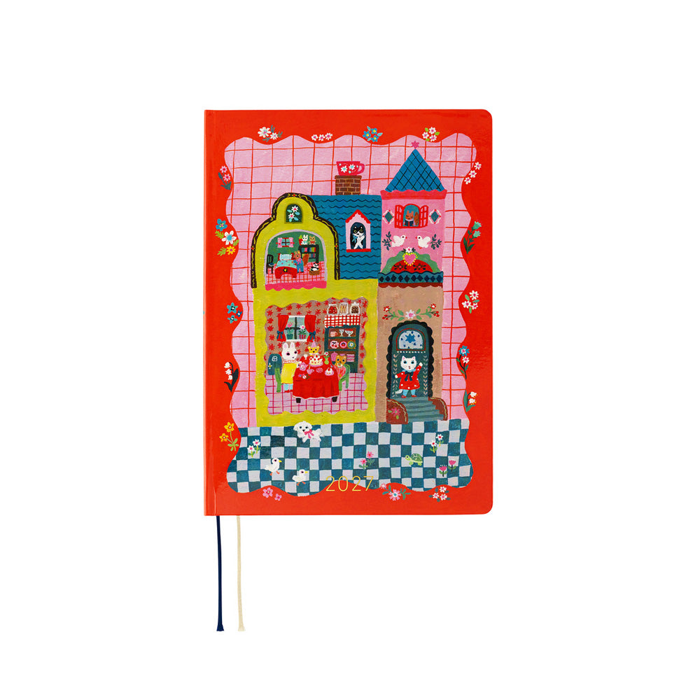 A hardcover notebook with a bright red cover featuring a whimsical, colorful patchwork-style illustration of houses and flowers. A small black and white ribbon bookmark hangs from the bottom left corner.